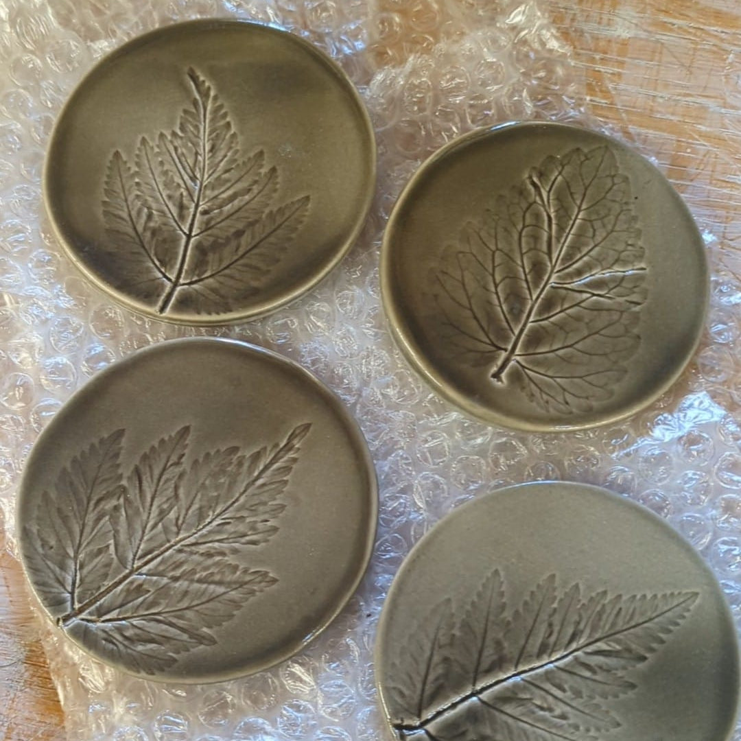 Four metallic coasters with leaf designs on a bubble wrap-covered surface.