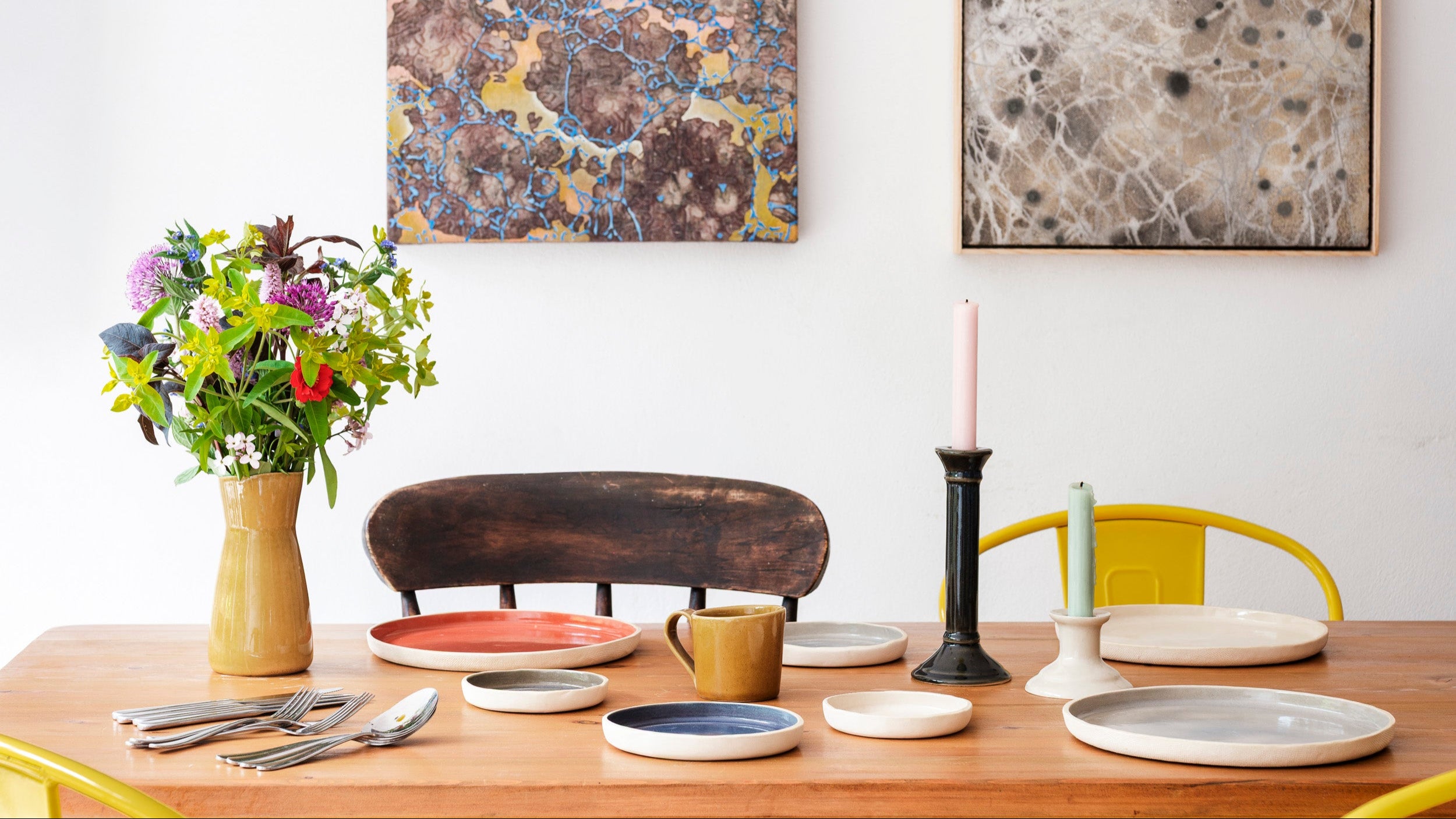 Dining table setting with plates, cups, and a vase of flowers, with abstract art and yellow pendant lights in the background.