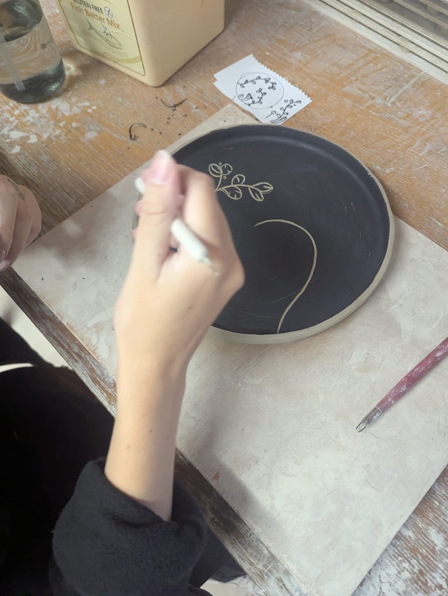 Person decorating a ceramic plate with a white tool on a wooden table. - the wirksworth pottery company