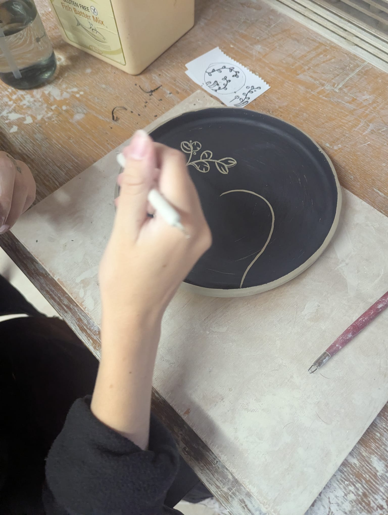 Person decorating a ceramic plate with a white tool on a wooden table. - the wirksworth pottery company